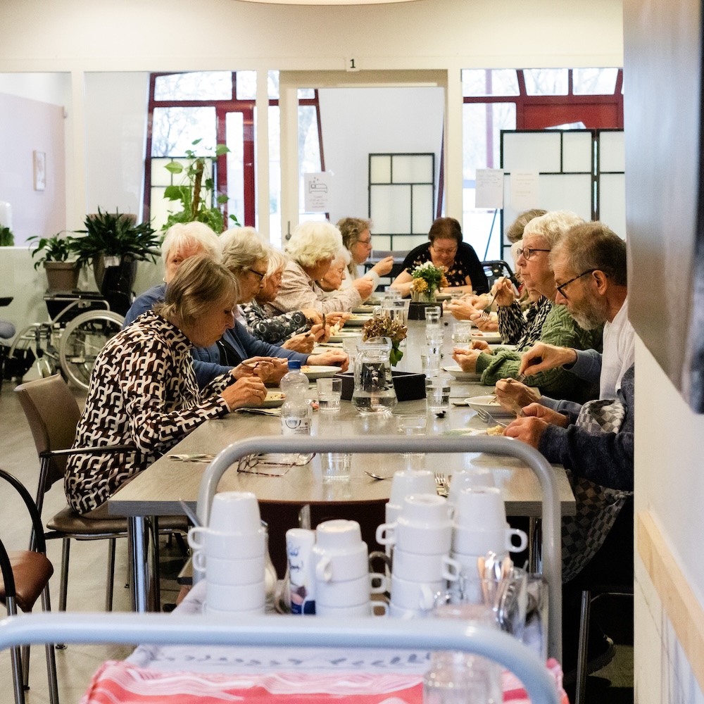 Bezoekers van de SOOS middag eten met elkaar aan een lange tafel