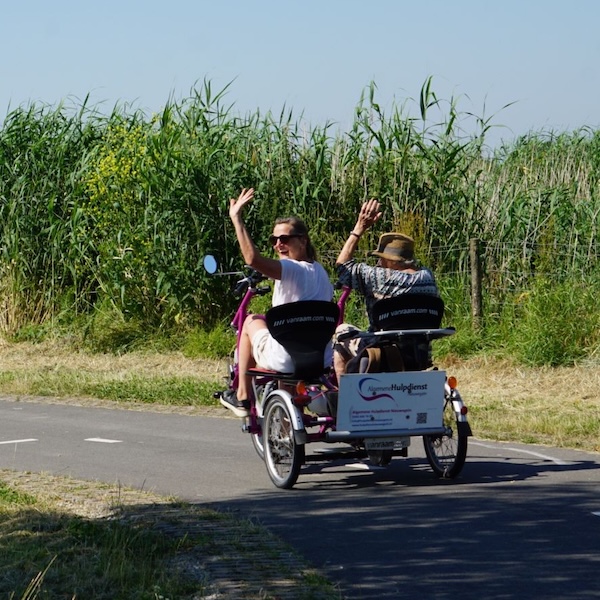 Vrijwilliger met hulpvrager fietsen op een duo fiets de natuur in en zwaaien achterom